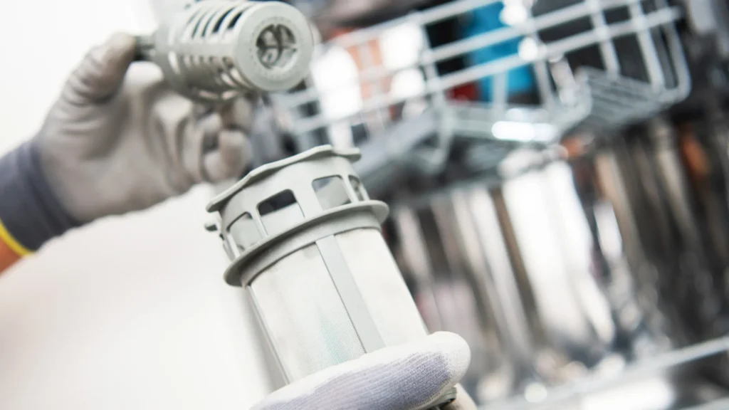 Gloved hands holding a cylindrical dishwasher filter above the open tub