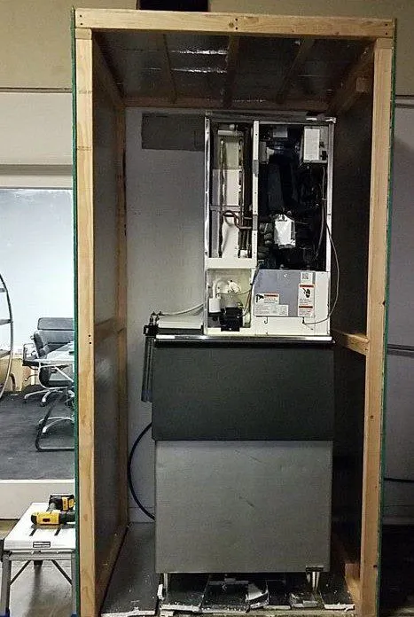 Ice machine with its front panels removed inside a close-fitting enclosure